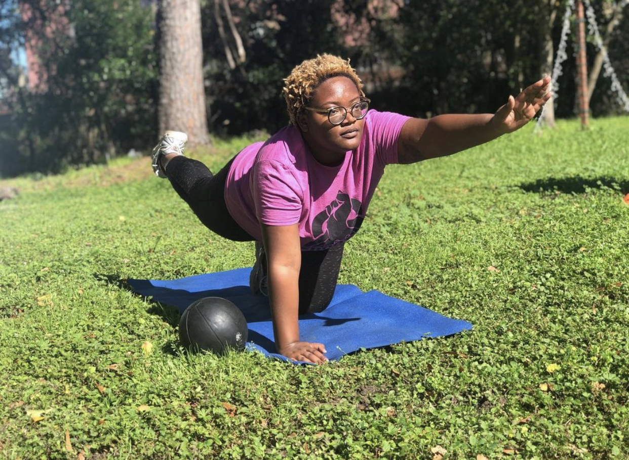 A rootcamp participant on a yoga mat on bright grass, extending an arm and opposite leg and looking forward