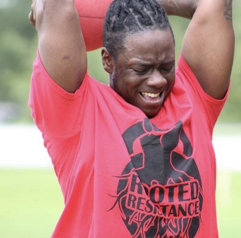 Maliq, a rootcamp participant in a red Rooted Resistance shirt slamming a medicine ball outdoors.